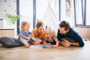 A family of four, mom, dad and two little kids, lies on a wood floor and looks at a book. There is a play tent in the background. 